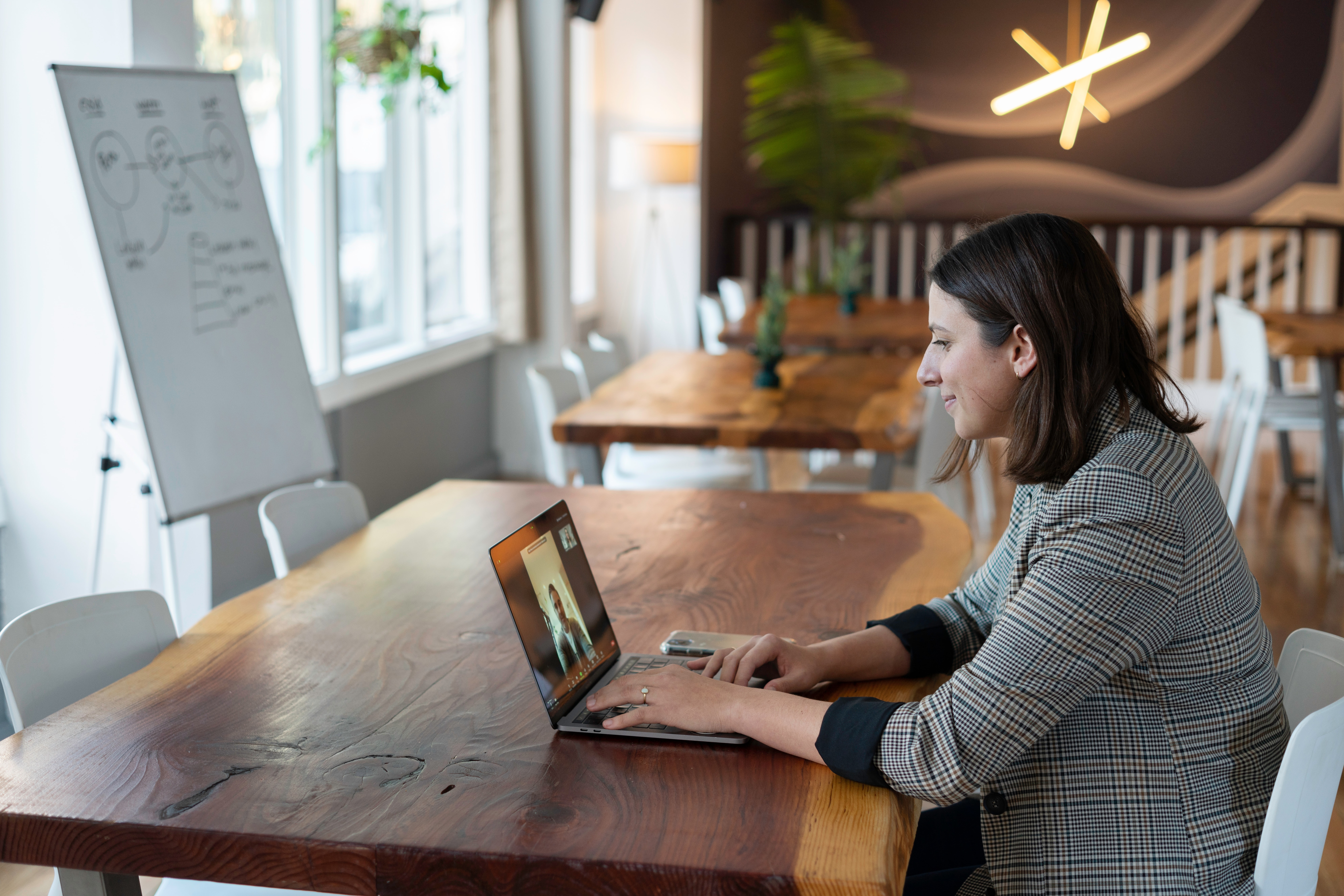 Uma mulher está sentada em uma mesa, em frente a um notebook. O espaço parece ser um café, mas ela está sozinha. Do lado esquerdo da foto podemos ver um quadro, com algumas coisas escritas. 
