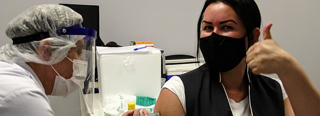 mulher morena, de máscara e com colete e crachá do MAckenzie, recebe injeção de outra mulher paramentada com máscara, jaleco e touca.