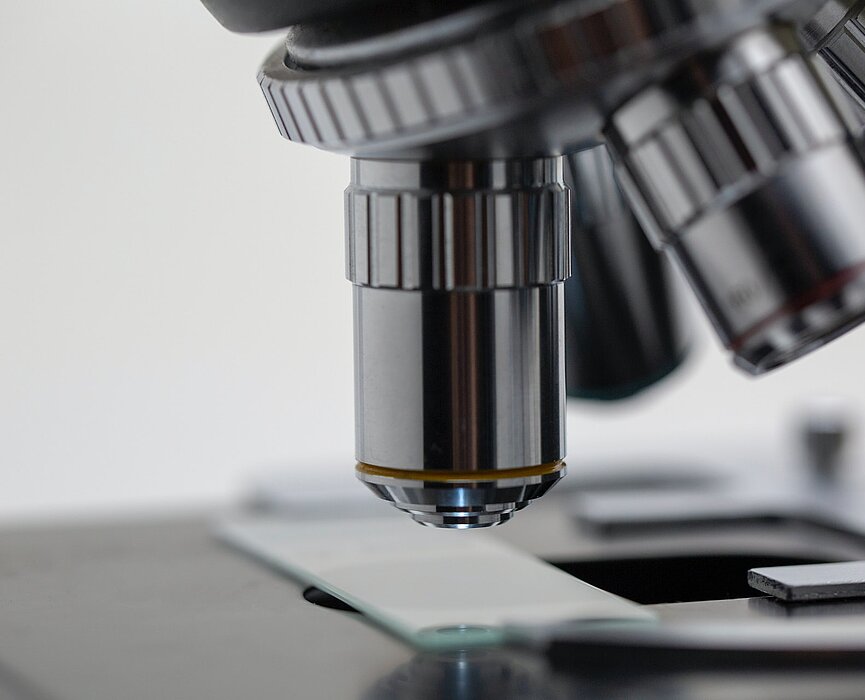 na imagem, uma foto bem próxima de uma das lentes de um microscópio nos tons cromado e prata analisando uma lâmina de vidro