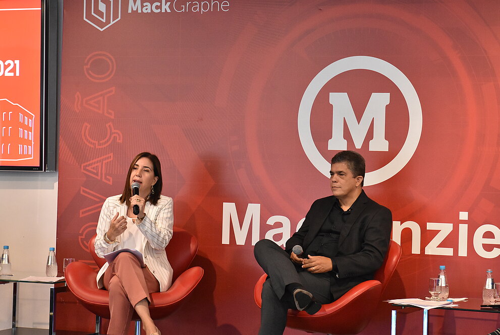 Janette e Marco Tullio estão sentados, em frente a um banner do Mackenzie.