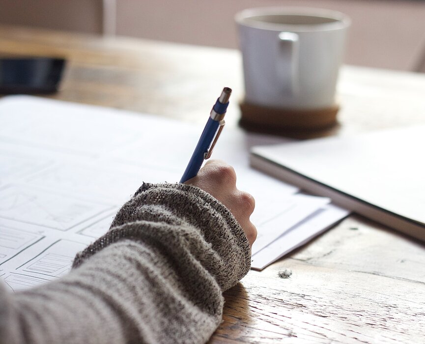 mão de garota segurando lápis e escrevendo em um caderno sobre uma mesa com uma xícara ao lado