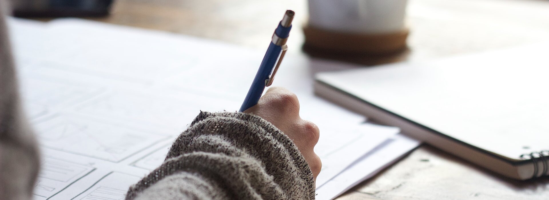 mão de garota segurando lápis e escrevendo em um caderno sobre uma mesa com uma xícara ao lado