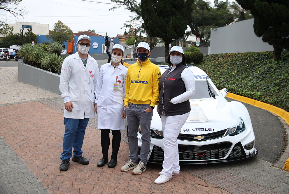 Quatro pessoas posam para foto ao lado do carro branco da Stock Car estacionado em frente ao HUEM.