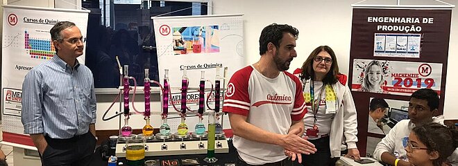 Pessoas de pé explicam sobre o curso aos visitantes sentados. Ao fundo há materiais de laboratório químico com diversas cores