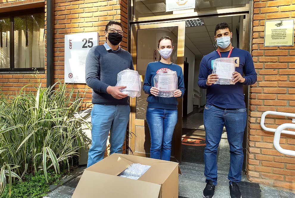 Três pessoas de pé, usando máscaras, em frente a uma parede de tijolos vermelhos seguram face shields 