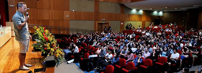 Thái à esquerda, sobre o palco, conversa com auditório cheio, à direita.
