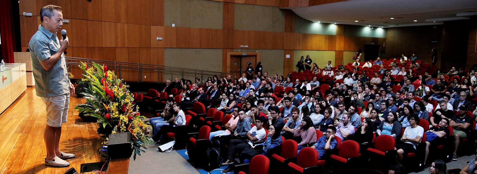 Thái à esquerda, sobre o palco, conversa com auditório cheio, à direita.