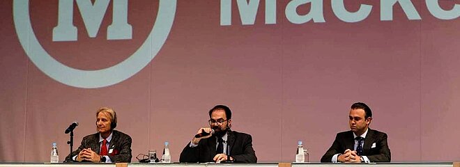 Palestrantes sentados à mesa, no palco, com tela com imagem do Mackenzie ao fundo.