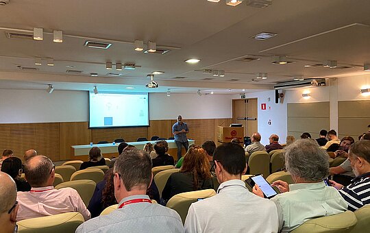Na imagem, um auditório lotado com uma imagem sendo apresentada por um rapaz no palco.