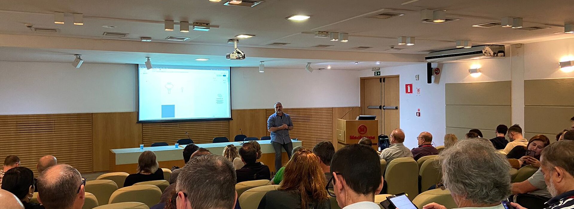 Na imagem, um auditório lotado com uma imagem sendo apresentada por um rapaz no palco.