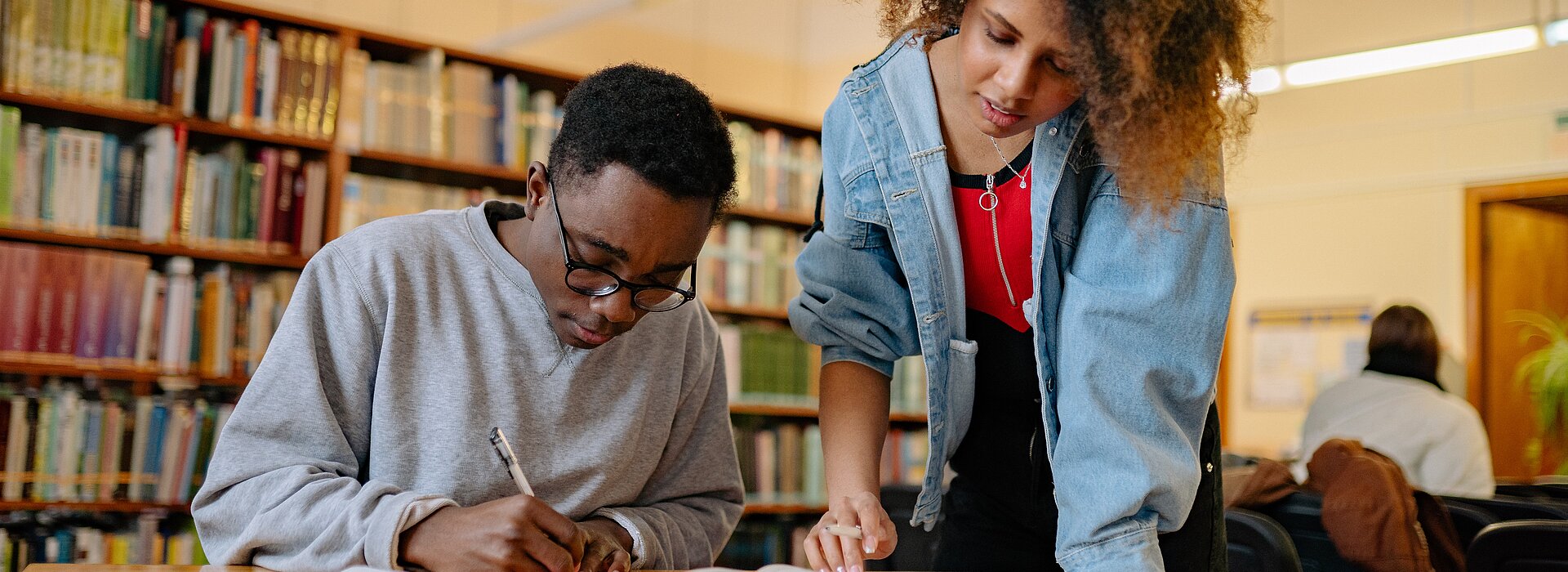 Um homem negro está sentado escrevendo e uma mulher negra está em pé olhando os materiais ao seu lado. Eles estão em uma biblioteca. 