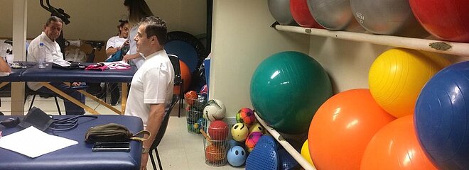 Do lado direito da imagem tem uma parede com bolas de ginástica de várias cores e do lado direito está um homem sentado fazendo exercícios ao mesmo tempo que outro homem também faz no fundo da imagem