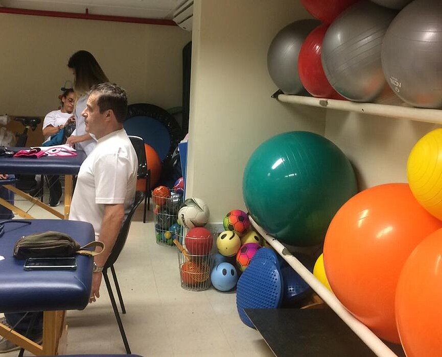 Do lado direito da imagem tem uma parede com bolas de ginástica de várias cores e do lado direito está um homem sentado fazendo exercícios ao mesmo tempo que outro homem também faz no fundo da imagem