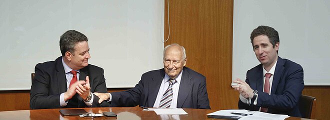 Três homens sentados ao redor de uma mesa redonda de madeira se cumprimentam.