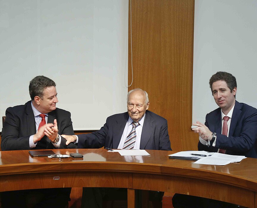 Três homens sentados ao redor de uma mesa redonda de madeira se cumprimentam.