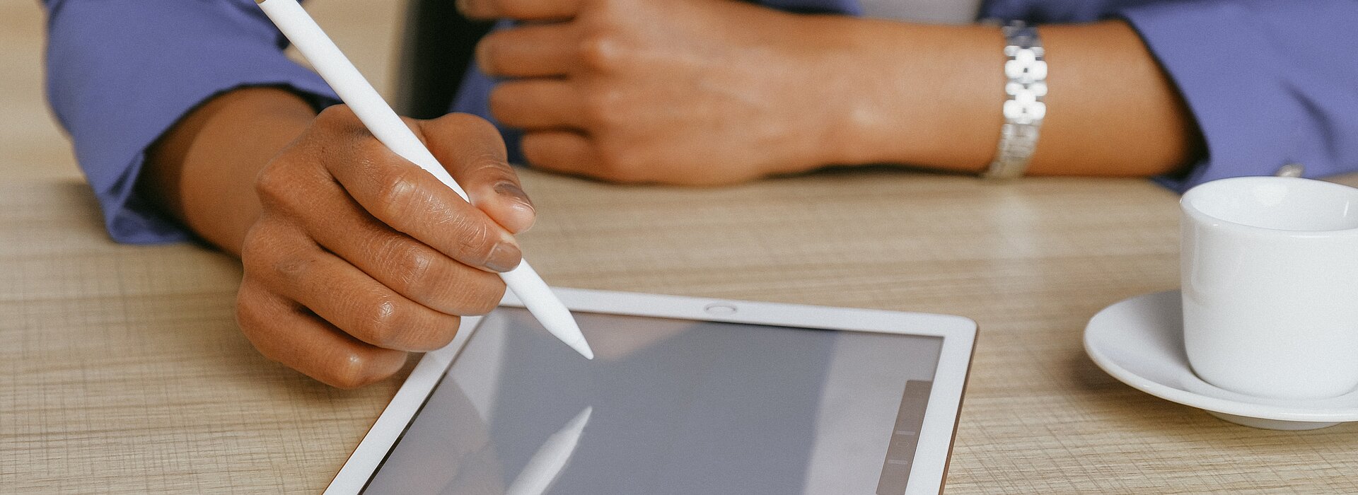 A foto mostra os braços de uma mulher apoiados sobre uma mesa. Ela está usando um tablet, com uma caneta. Uma xícara também está ao seu lado. Ela está usando um blazer, o que remete à negócios.