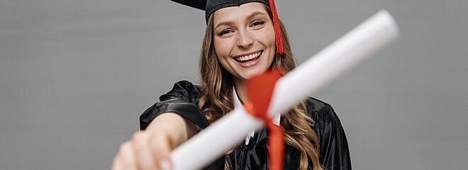 Na foto, uma menina mostra seu diploma para a câmera. Ela está de beca, com capelo e está sorrindo.
