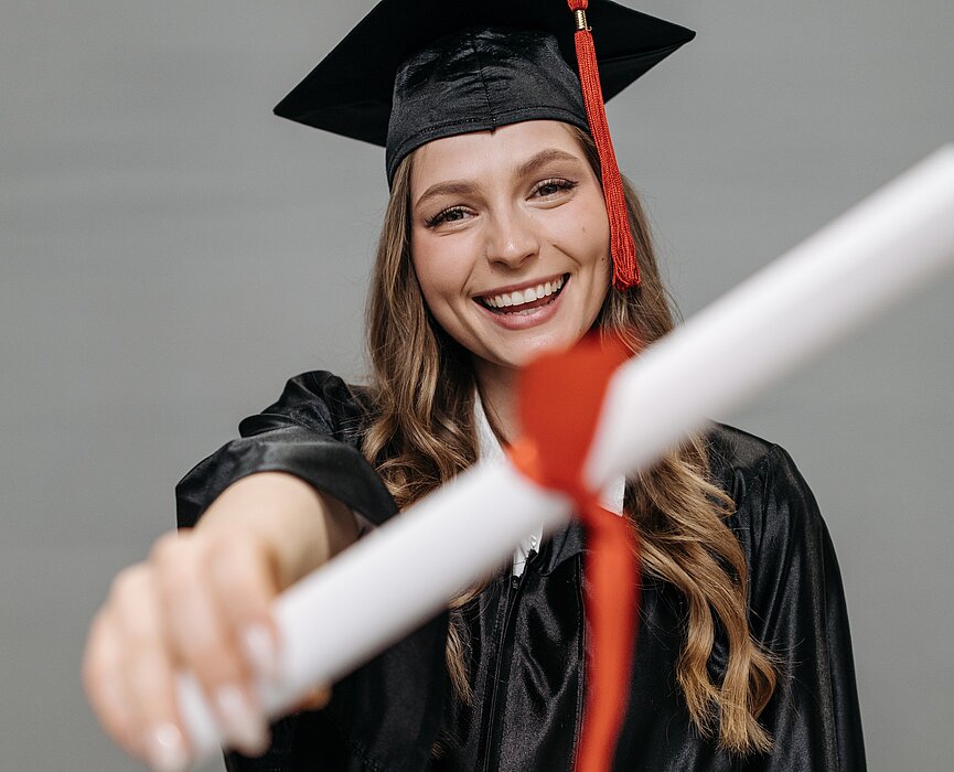 Na foto, uma menina mostra seu diploma para a câmera. Ela está de beca, com capelo e está sorrindo.