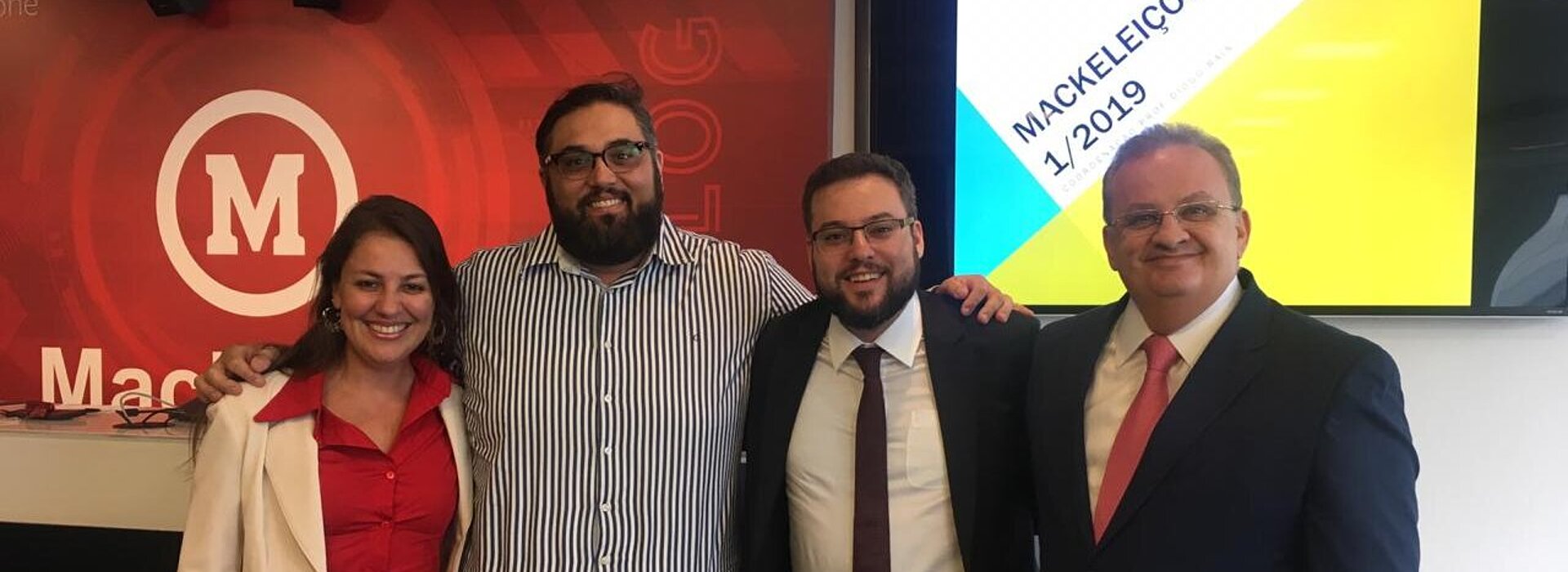 Pesquisadores do evento posando para a foto abraçados em pé em frente à projeção da escrita: "Mackeleições". 