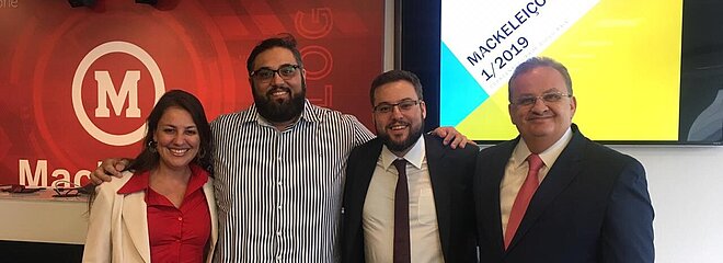 Pesquisadores do evento posando para a foto abraçados em pé em frente à projeção da escrita: "Mackeleições". 