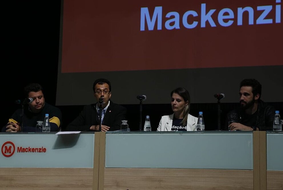 Palestrantes sentados em frente a mesa voltada para público