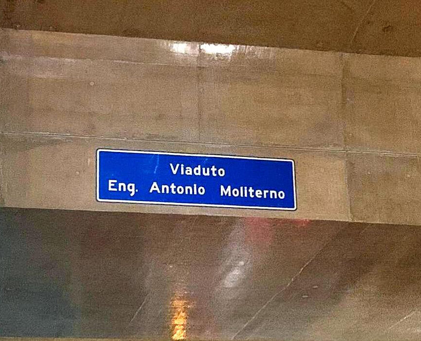 Na imagem, é possível ver a lateral de um viaduto na cor cinza concreto com uma placa de fundo azul com letras brancas onde é possível ler "Engenheiro Antonio Moliterno"