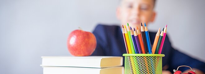 Imagem contém livros, uma maça, um relógio, um óculos, um recipiente com lápis e um menino no fundo.