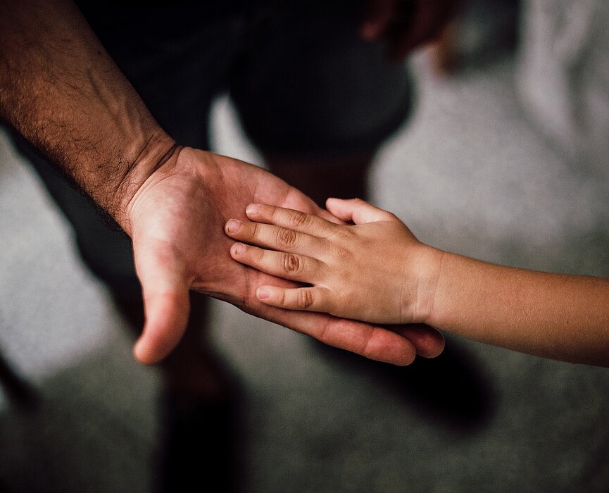 Um adulto estica a mão para uma criança. A foto tem foco na mão deles. 