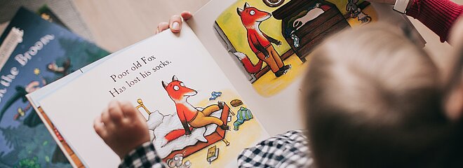 A foto foi tirada de cima e mostra a cabeça de uma criança e suas mãos em cima de um livro, com ilustrações de uma raposa. A criança está acompanhando por um adulto, porque conseguimos ver a mão de uma mulher adulta, ajudando a criança na leitura.
