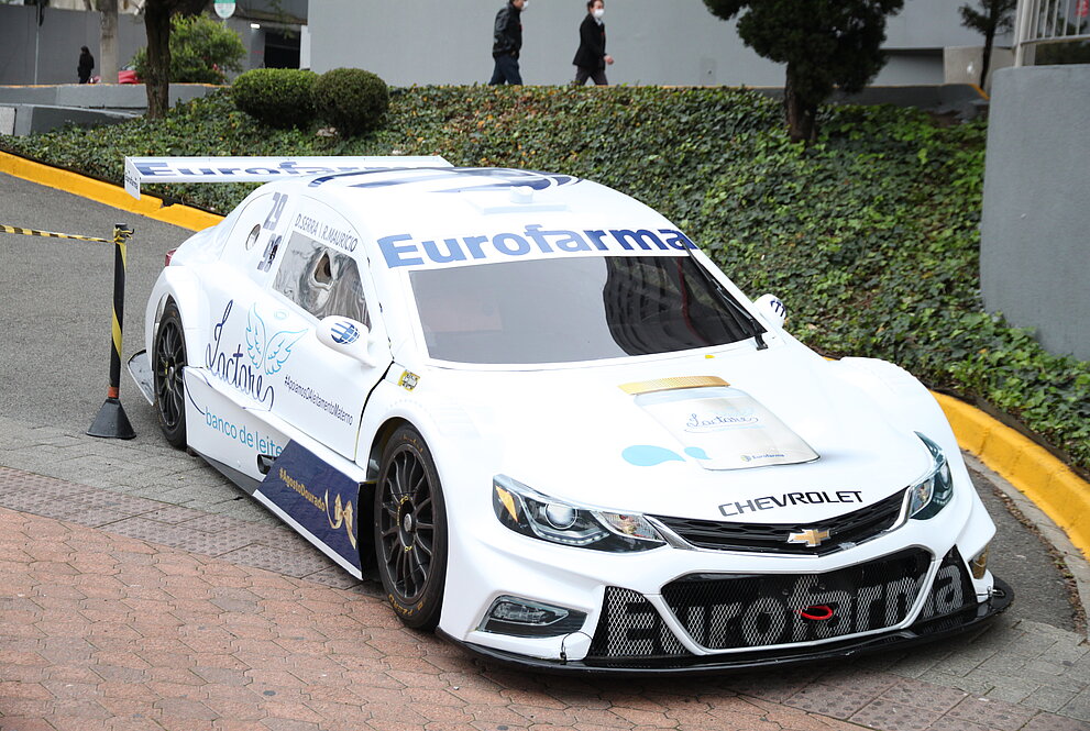 Carro branco da Stock Car estacionado em frente ao HUEM. Atrás pode-se ver o prédio cinza  e um jardim