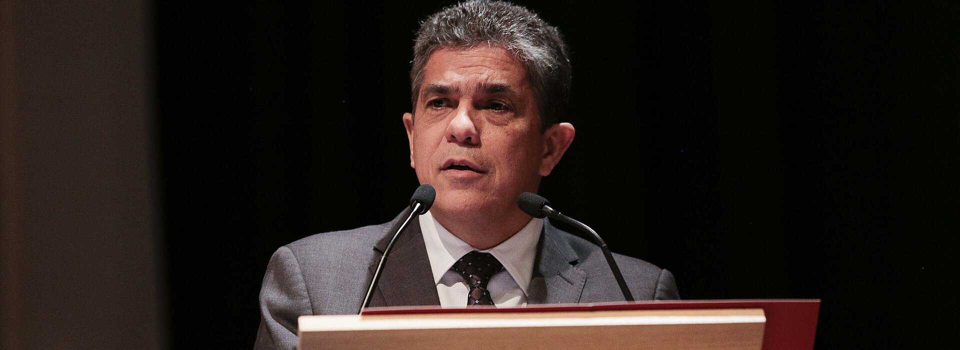 Na foto, Marco Tullio, reitor da Universidade, está falando em um microfone atrás de um púlpito. A foto está bem focada em seu rosto. Ele está usando terno e olhando para a lateral esquerda.
