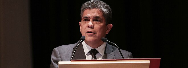 Na foto, Marco Tullio, reitor da Universidade, está falando em um microfone atrás de um púlpito. A foto está bem focada em seu rosto. Ele está usando terno e olhando para a lateral esquerda.
