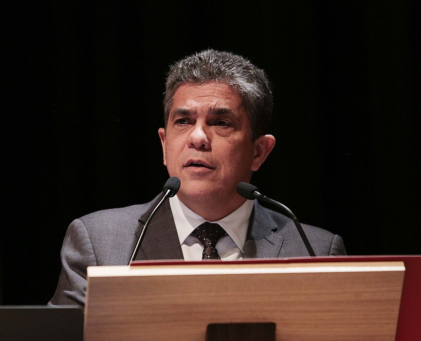 Na foto, Marco Tullio, reitor da Universidade, está falando em um microfone atrás de um púlpito. A foto está bem focada em seu rosto. Ele está usando terno e olhando para a lateral esquerda.
