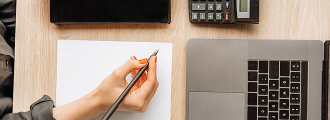 Foto foi tirada de cima e mostra uma mão, segurando uma caneta e escrevendo em um papel. Ao seu lado tem um caderno, calculadora e um notebook.
