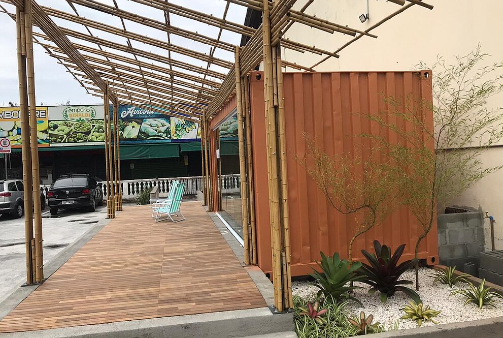 A foto mostra a lateral da cozinha comunitária. Um pequeno jardim fica em torno da cozinha construída. Logo na frente, duas cadeiras de praia ficam no espaço aberto da cozinha.
