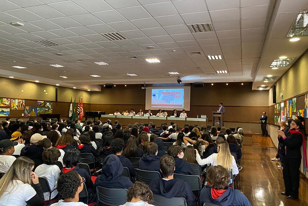 estudantes para debate reunidos na mesa do auditório com telão ao fundo e plateia cheia de estudantes sentados