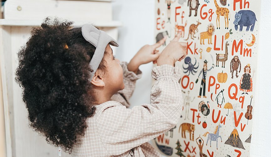 Uma menina de aproximadamente quatro anos está em frente a um painel cheio de letras do alfabeto. Todas as letras têm ao lado um animal, como girafa, elefante.