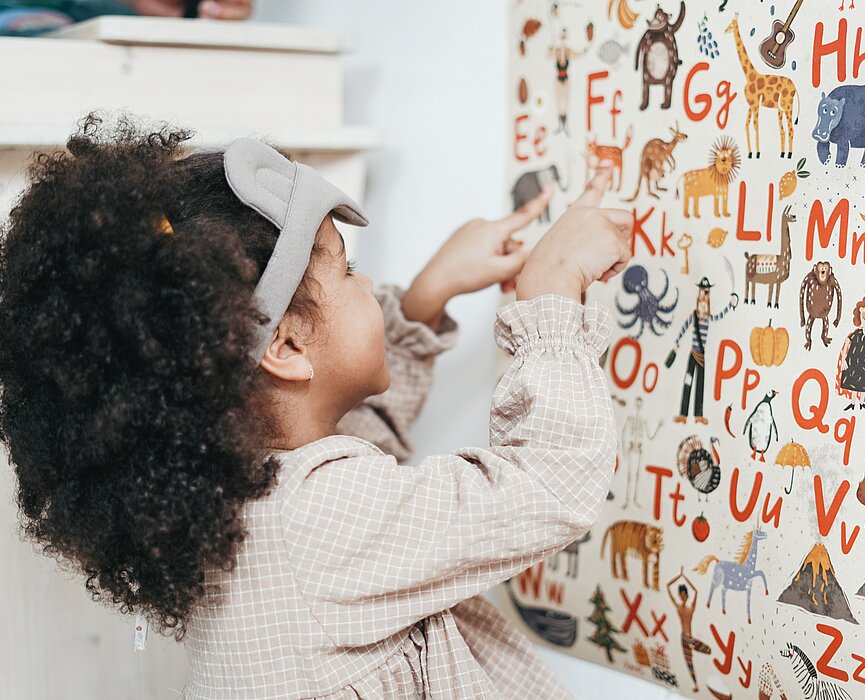 Uma menina de aproximadamente quatro anos está em frente a um painel cheio de letras do alfabeto. Todas as letras têm ao lado um animal, como girafa, elefante. 