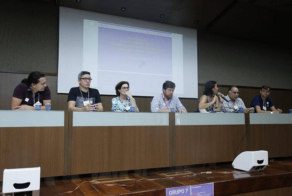 Palestrantes sentados em mesa no palco. Telão com slides na parte de trás da imagem