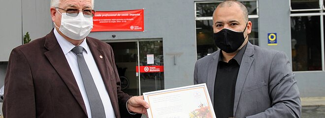 Dois homens com máscaras e ternos seguram um quadro de reconhecimento em frente ao hospital
