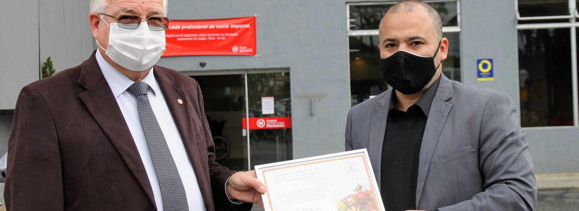 Dois homens com máscaras e ternos seguram um quadro de reconhecimento em frente ao hospital