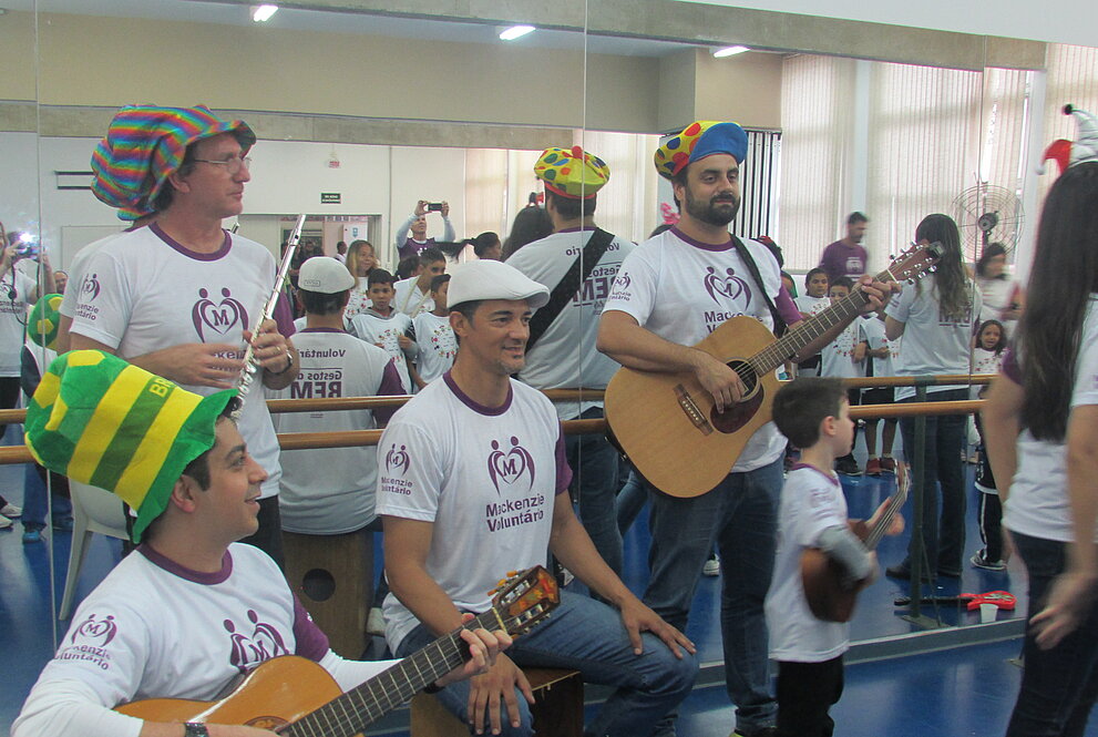 voluntários estão com violão e instrumentos de percussão na mão enquanto animam o ambiente. Todos estão com chapéus coloridos. 