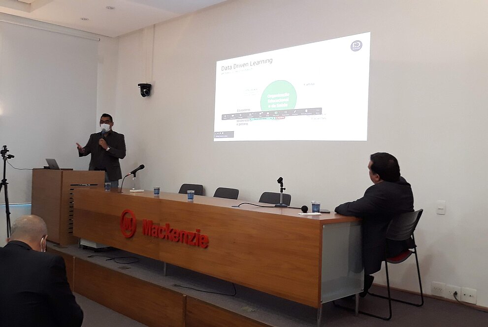 um homem de pé, utilizando terno e máscara, apresenta slides. Há uma mesa de madeira, com outro homem sentado. Pode-se ler o nome Mackenzie gravado na mesa em vermelho.