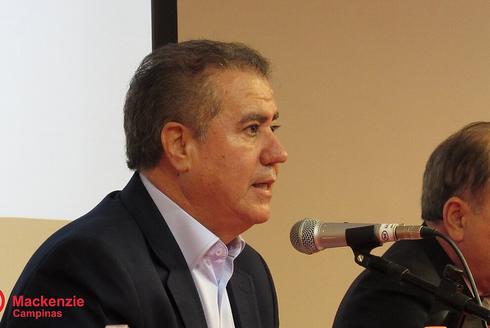 homem sentado discursa ao microfone na mesa de autoridades
