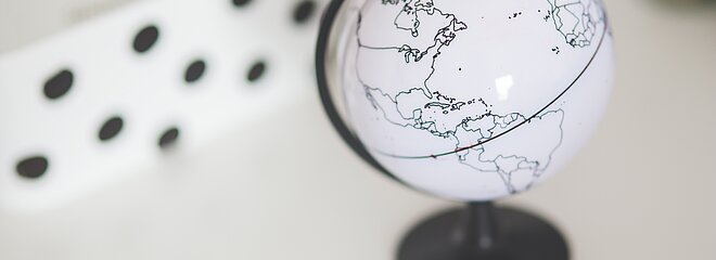 A imagem mostra um globo do mundo ao lado direito. No fundo da imagem, aparece uns detalhes em preto por cima de uma parte branca, lembrando uma peça de dominó. 