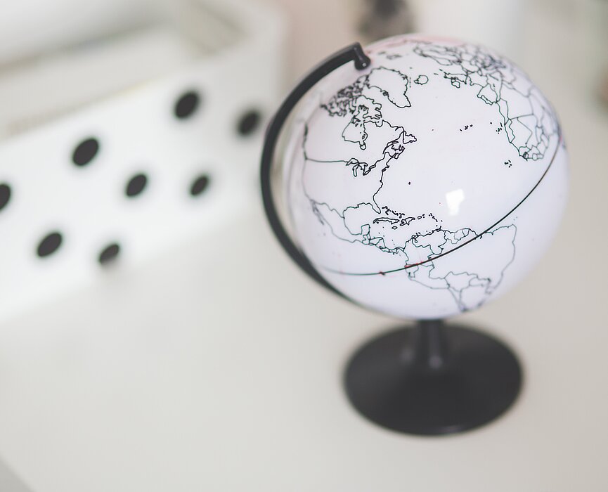 A imagem mostra um globo do mundo ao lado direito. No fundo da imagem, aparece uns detalhes em preto por cima de uma parte branca, lembrando uma peça de dominó. 