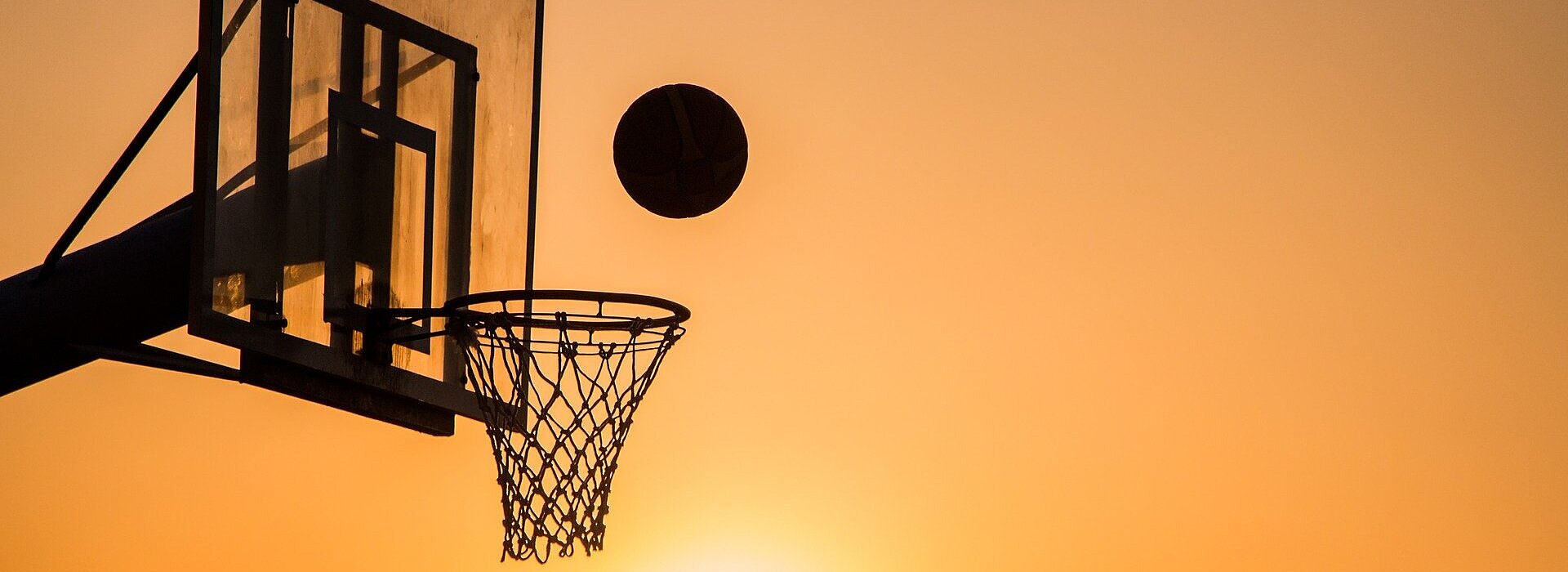 Imagem mostra uma cesta de basquete com um homem jogando a bola em direção a ela.
