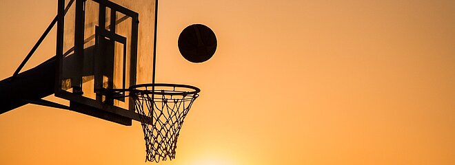 Imagem mostra uma cesta de basquete com um homem jogando a bola em direção a ela.