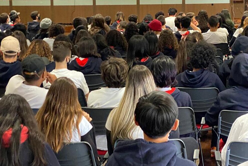 estudantes para debate reunidos na mesa do auditório com telão ao fundo e plateia cheia de estudantes sentados