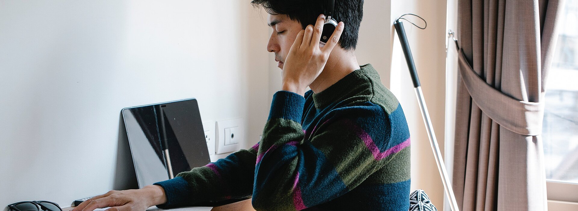 A foto mostra um homem sentado em frente a um computador fazendo a leitura em braile de um livro com sua bengala vermelha atrás